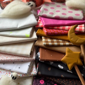 May include: A collection of folded fabric squares featuring various patterns including pink polka dots, red and white gingham, brown plaid, and black prints with ghosts, pumpkins, and spiderwebs, accompanied by a wooden star and moon wand.