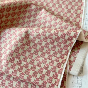 Puede incluir: Tela de color crema con un patrón repetido de motivos florales rosas. La tela está doblada, revelando la textura y el borde sin rematar del material. Ideal para proyectos de costura, la tela tiene un aspecto suave y acogedor.