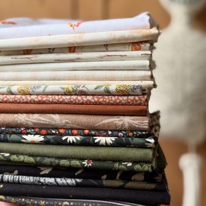 May include: A stack of folded fabric pieces featuring a variety of patterns and solid colors. The collection includes white, cream, and beige prints, followed by floral, solid rust, brown, dark green, and black fabrics with celestial and botanical motifs.