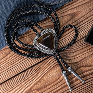 Op de afbeelding: Een zwarte leren bolo tie met een driehoekige zilveren hanger met een bruine tijgeroog-steen.
