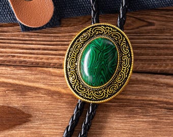 May include: Oval bolo tie with a green stone center and a gold-colored filigree border. The tie features a black braided cord and a brown leather strap. The background is a wooden surface.