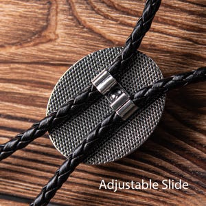 May include: A black bolo tie with an adjustable slide. The tie has a braided leather cord and a round metal pendant.