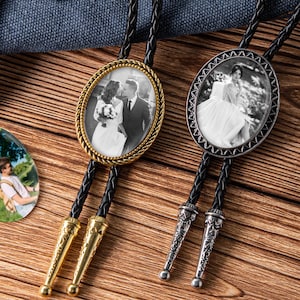 May include: Two bolo ties with oval photo pendants. One has a gold-toned frame with a braided leather cord and gold-toned tips. The other has a silver-toned frame, a braided leather cord, and silver-toned tips. Both pendants feature black and white wedding photos.