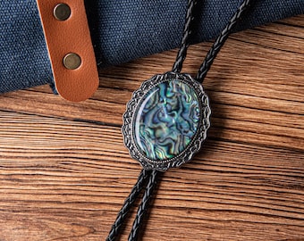 May include: Oval bolo tie with a silver-toned filigree frame and an iridescent abalone shell center. The black braided leather cord is visible against a wooden surface. A brown leather strap with two brass-colored rivets is in the background.