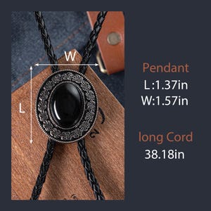 May include: A black bolo tie with a silver-toned pendant featuring a black oval stone surrounded by an intricate design. The pendant measures 1.37 inches long and 1.57 inches wide. The cord is 38.18 inches long.