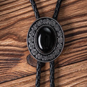 May include: A black bolo tie with a silver oval pendant featuring an intricate design and a black gemstone.