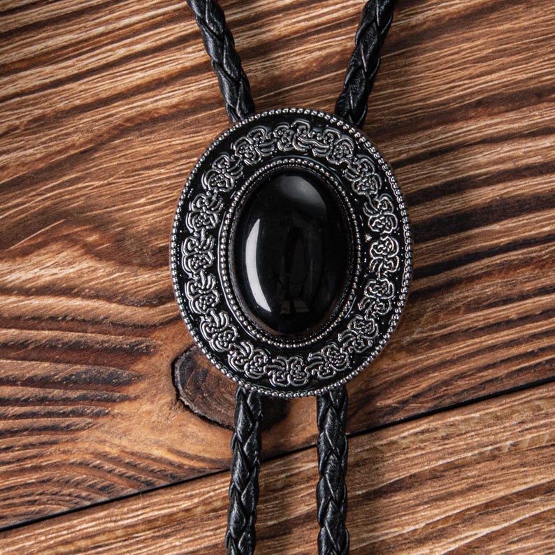 May include: A black bolo tie with a silver oval pendant featuring an intricate design and a black gemstone.