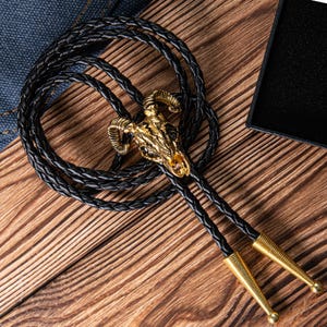 May include: A black leather bolo tie with a gold-toned ram skull pendant. The tie is braided and has two long tassels.