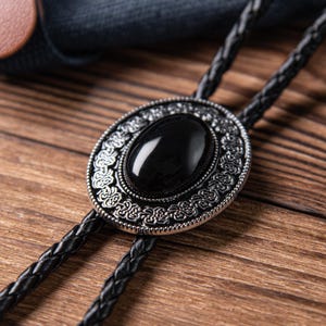 May include: A black bolo tie with a silver oval pendant featuring an intricate design and a black gemstone in the center.