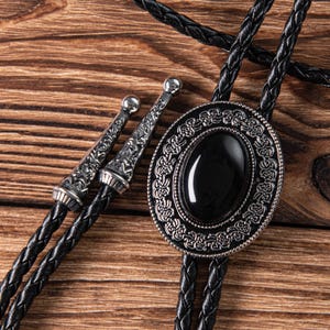May include: A black bolo tie with a silver oval pendant featuring a black gemstone surrounded by an intricate design.