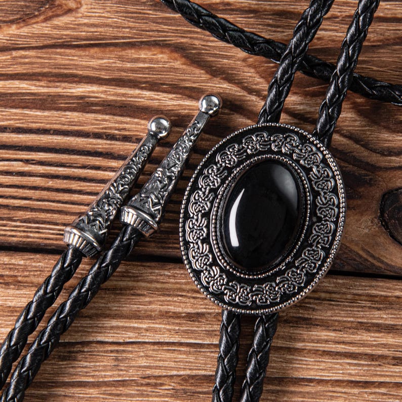 May include: A black bolo tie with a silver oval pendant featuring a black gemstone surrounded by an intricate design.