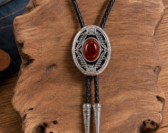 May include: A bolo tie featuring a central oval red stone set in an intricate silver-tone metal frame with filigree details. The cord is made of braided black leather and terminates in two matching silver-tone metal tips with engraved patterns.