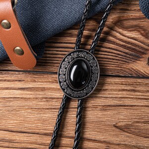 May include: A black bolo tie with a silver-toned oval pendant featuring a black gemstone. The pendant has an intricate design with a floral pattern.