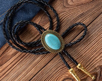 May include: A black braided leather bolo tie with a turquoise oval stone centerpiece set in a gold-colored filigree frame. The tie features gold-colored metal tips and is displayed on a wooden surface.