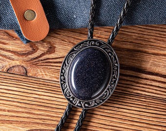 May include: A bolo tie featuring a dark blue, oval-shaped stone with a silver-colored, ornate border. The tie has a braided black cord and a brown leather strap with two metal rivets.