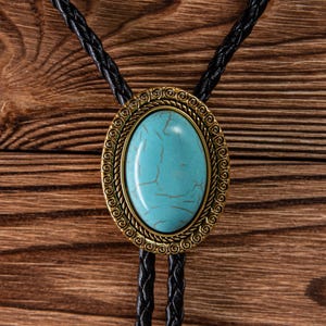 May include: Oval turquoise bolo tie with a gold-colored filigree frame and black braided leather cord. The turquoise stone has brown veining. The bolo tie is a western-style accessory.