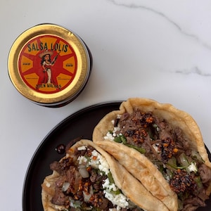 May include: Two tacos filled with meat, cheese, and cilantro on a black plate. A jar of salsa labeled "Salsa Lolis Bien Macha" is in the background.