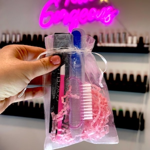 Può includere: Un piccolo sacchetto di rete bianca riempito con carta velina rosa e strumenti per la cura delle unghie. Gli strumenti includono una lima per unghie nera, una lima per unghie rosa, una lima per unghie trasparente e uno spazzolino per unghie bianco. Il sacchetto è tenuto in una mano davanti a un muro con un cartello al neon rosa che dice "Hello Gorgeous".
