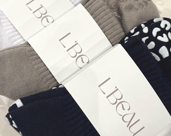 Op de afbeelding: Drie paar geribbelde sokken in wit, beige en zwart, elk met een luipaardprint en verhoogde siliconen noppen op de zolen. Elk paar is bevestigd met een witte papieren band waarop de merknaam L'Beau in zwarte letters is gedrukt.