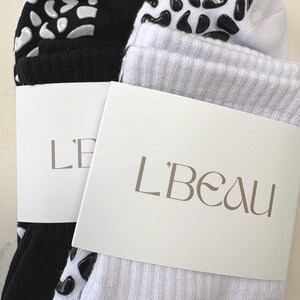 Könnte beinhalten: Ein Paar gerippte Socken, eine in Schwarz und eine in Weiß, mit einem rutschfesten Leopardenmuster auf den Sohlen. Jede Socke ist mit einer weißen Papierbanderole versehen, auf der der Markenname L'BEAU in einer eleganten Serifenschrift steht.