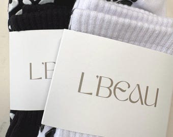 Op de afbeelding: Een paar geribbelde sokken, één zwart en één wit, met een antislip luipaardpatroon op de zolen. Elke sok is verpakt in een witte papieren band waarop de merknaam L'BEAU in een elegant schreeflettertype is gedrukt.