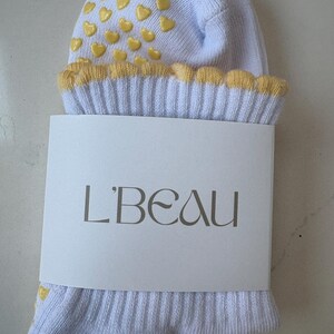 May include: A pair of light blue socks with yellow heart-shaped grips on the top. The cuff is trimmed with yellow scalloped edges. A white label with the text "L'BEAU" is wrapped around the socks.