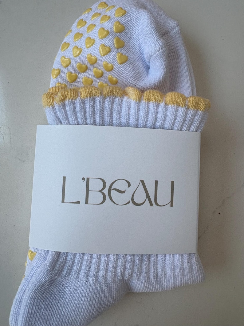 May include: A pair of light blue socks with yellow heart-shaped grips on the top. The cuff is trimmed with yellow scalloped edges. A white label with the text "L'BEAU" is wrapped around the socks.