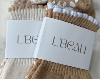 Op de afbeelding: Twee paar sokken, één beige en één bruin, elk verpakt met een witte band met de tekst "L'Beau". De beige sokken hebben hartvormige gripjes, terwijl de bruine sokken witte luipaardprint gripjes hebben. Beide paren hebben een witte gefranjerde rand.