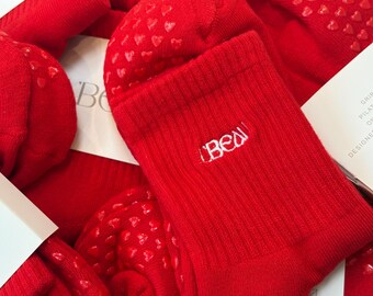 Op de afbeelding: Felrode sokken van kuitlengte met een geribbelde boord, de geborduurde witte tekst "BEAU" en een patroon van kleine rode hartvormige siliconen grippers op de zolen. De sokken worden getoond naast witte productinformatiekaarten.