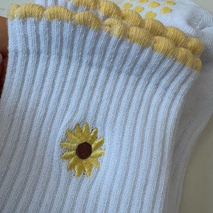 May include: White ribbed socks with a yellow sunflower embroidery and a scalloped yellow trim. The socks have yellow heart-shaped grips on the sole. The sunflower has a brown center and yellow petals.