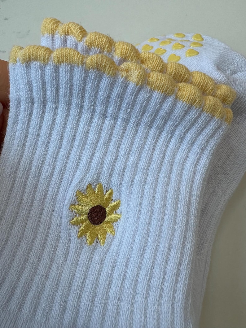 May include: White ribbed socks with a yellow sunflower embroidery and a scalloped yellow trim. The socks have yellow heart-shaped grips on the sole. The sunflower has a brown center and yellow petals.