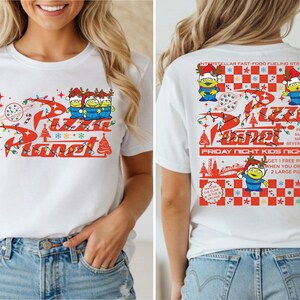 A woman wearing a white t-shirt with a red and white design featuring a cartoon character and text related to a pizza-themed event.