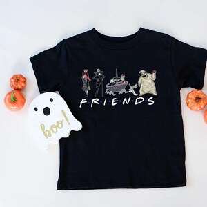 A black t-shirt with a graphic design featuring characters from the TV show Friends, including Jack Skellington, Mary Jane, and Oogie Boogie, along with the text "FRIENDS". The t-shirt is placed on a white surface, and there are small orange pumpkins arranged around it.
