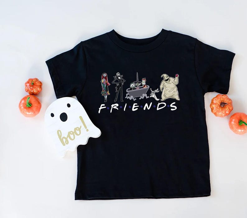 A black t-shirt with a graphic design featuring characters from the TV show Friends, including Jack Skellington, Mary Jane, and Oogie Boogie, along with the text "FRIENDS". The t-shirt is placed on a white surface, and there are small orange pumpkins arranged around it.