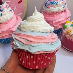 Puede incluir: Tres cupcakes rosas y blancos con glaseado blanco y chispas. Los cupcakes están decorados con una cereza roja en la parte superior. Un cupcake está siendo sostenido en una mano.
