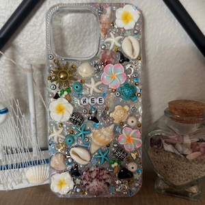 May include: Clear phone case decorated with seashells, starfish, flowers, and beads. The name "KEE" is spelled out in letter beads. The case has a glittery background and a variety of colors, including white, pink, blue, and gold.