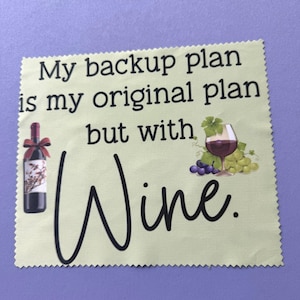 May include: A square, light yellow microfiber cleaning cloth with pinked edges. It features the black text, My backup plan is my original plan but with Wine. An illustration of a wine bottle with a red bow and a glass of red wine with grapes.
