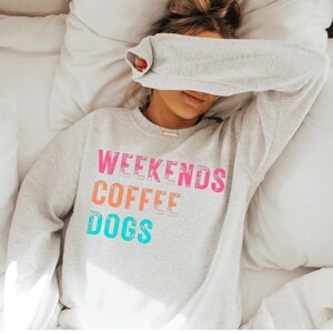 Może przedstawiać: Jasnoszara bluza z napisem "WEEKENDS COFFEE DOGS" w kolorze różowym, pomarańczowym i turkusowym. Osoba nosząca bluzę leży na łóżku z białymi prześcieradłami i poduszkami.