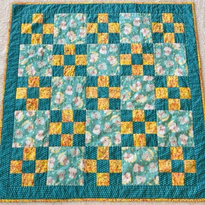 May include: A handmade baby quilt with a patchwork design. The quilt features a teal border with white polka dots and a yellow edge. The main body has alternating squares of yellow and teal fabric with a sheep and moon pattern.
