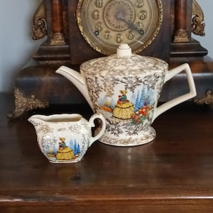 Könnte beinhalten: Ein Vintage-Teeservice mit einer Teekanne und einem Sahnekännchen. Das Set ist weiß mit goldenen Blumenmustern und einer farbenfrohen Illustration einer Frau in einem gelben Kleid. Die Teekanne hat einen Deckel und einen gebogenen Ausguss. Das Sahnekännchen hat einen Griff.