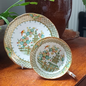 May include: A vintage teacup and saucer set. The white porcelain cup and saucer feature a gold rim and a colorful floral and bird design. The set is placed on a wooden surface, with a blurred background.