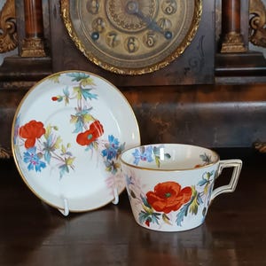 Può includere: Un set di tazza e piattino bianchi con un motivo floreale con papaveri rossi e fiori blu. La tazza e il piattino hanno finiture dorate. Il set è posto davanti a un orologio d'epoca.