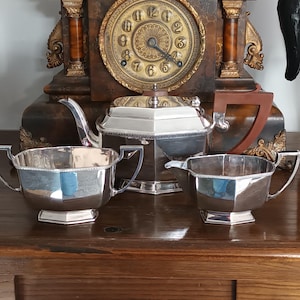 May include: A three-piece silver-plated tea set featuring an octagonal teapot with a brown handle, a matching sugar bowl with two handles, and a creamer. Each piece has a polished metallic finish and a distinct geometric, multi-sided shape.