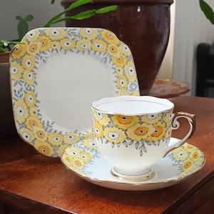 May include: A vintage teacup, saucer, and square plate set. The white porcelain pieces are decorated with a yellow floral pattern with grey accents. The teacup has a gold handle and trim. The set is displayed on a wooden surface.