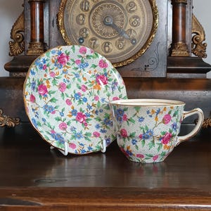 Puede incluir: Un juego de taza y platillo de té vintage con un estampado floral en rosa, azul y amarillo sobre un fondo crema. La taza y el platillo tienen ribetes dorados. El juego se exhibe sobre una superficie de madera oscura.
