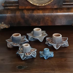 Könnte beinhalten: Fünf Kerzenhalter aus Keramik. Jeder Halter hat eine blütenförmige Basis in Braun- und Blau-Grau-Tönen mit einer zentralen zylindrischen Tasse. Die Kerzenhalter sind auf einer dunklen Holzoberfläche angeordnet.