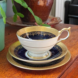 Op de afbeelding: Een vintage theeservies bestaande uit een kopje, schotel en bord. Het kopje en de schotel hebben een marineblauw interieur en midden met gouden filigraan en rand. De set staat op een houten oppervlak.