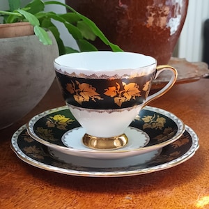 Könnte beinhalten: Ein Vintage-Tee-Set mit Tasse, Untertasse und Teller. Die Tasse hat einen weißen Innenraum und ein schwarzes Außenband mit goldenen Blattmustern. Untertasse und Teller haben passende Schwarz-Gold-Muster. Das Set steht auf einer Holzoberfläche.