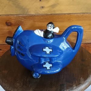Peut inclure: Une théière fantaisiste bleu cobalt en forme de biplan, avec une hélice noire et un pilote dans le cockpit. La théière porte des croix blanches sur les ailes et le texte "Blue Max". Elle est posée sur un tabouret en bois.
