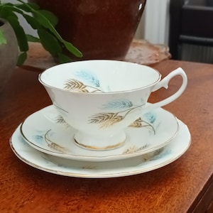 May include: A white teacup and saucer set with a matching side plate. The set features a gold trim and a delicate pattern of blue and gold leaves. The teacup has a curved handle.